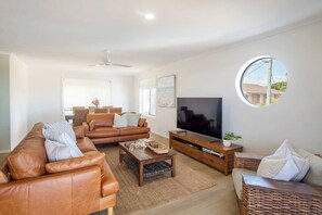 A modern living room with a brown leather sofa, a wooden coffee table, a TV on a wooden stand, and a round window with a view of the outside. - Crest on Convent - Oceanstays - Steps to Cafes, Beaches & Town (Yamba)