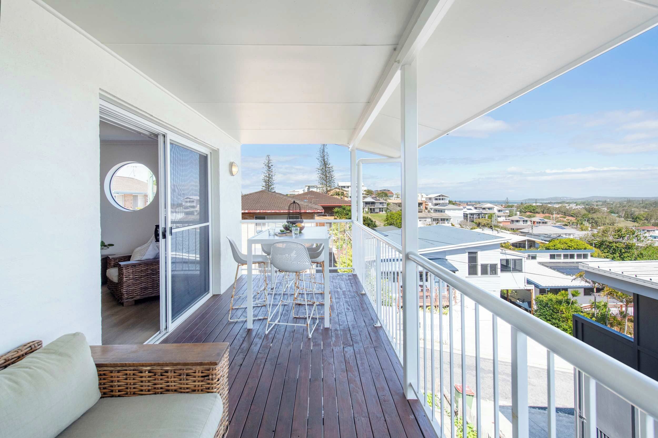 Uma varanda com piso de madeira, guarda-corpo branco e vista para o bairro. Há uma mesa com cadeiras e um sofá de vime.