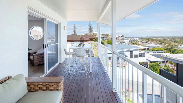 Ein Balkon mit Holzboden, weißer Reling und Blick auf die Nachbarschaft. Ein Tisch mit Stühlen und eine Rattan-Couch sind ebenfalls vorhanden.