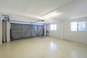 A clean, empty garage with a metal garage door, fluorescent lighting, and a window on the right wall. - Crest on Convent - Oceanstays - Steps to Cafes, Beaches & Town (Yamba)