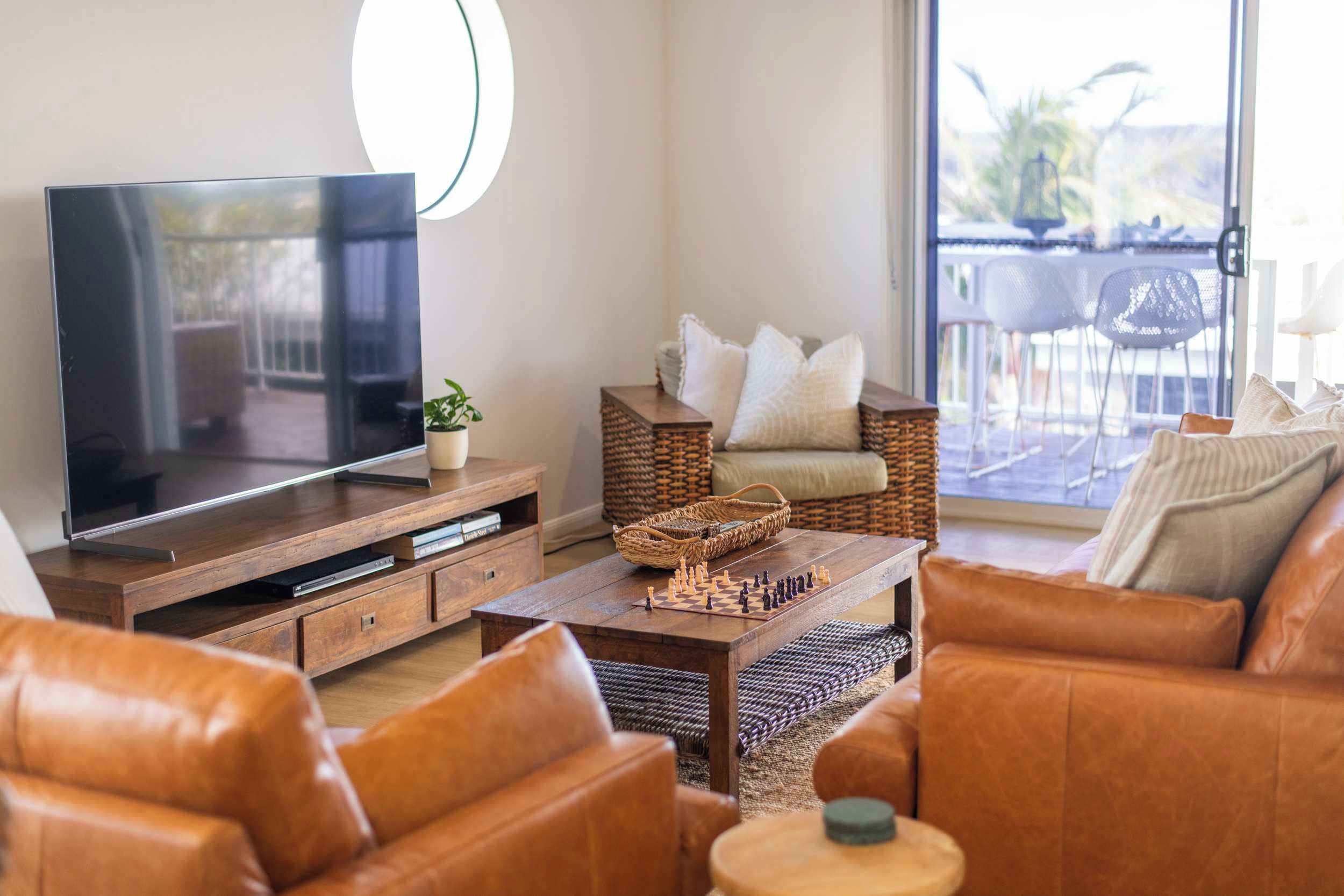 A living room with a flat-screen TV, leather sofas, a coffee table with a chess set, wicker furniture, and a view of the outdoors.