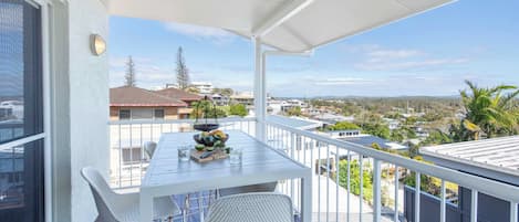 A balcony with a white table and chairs, a bowl of fruit, and a view of a residential area.