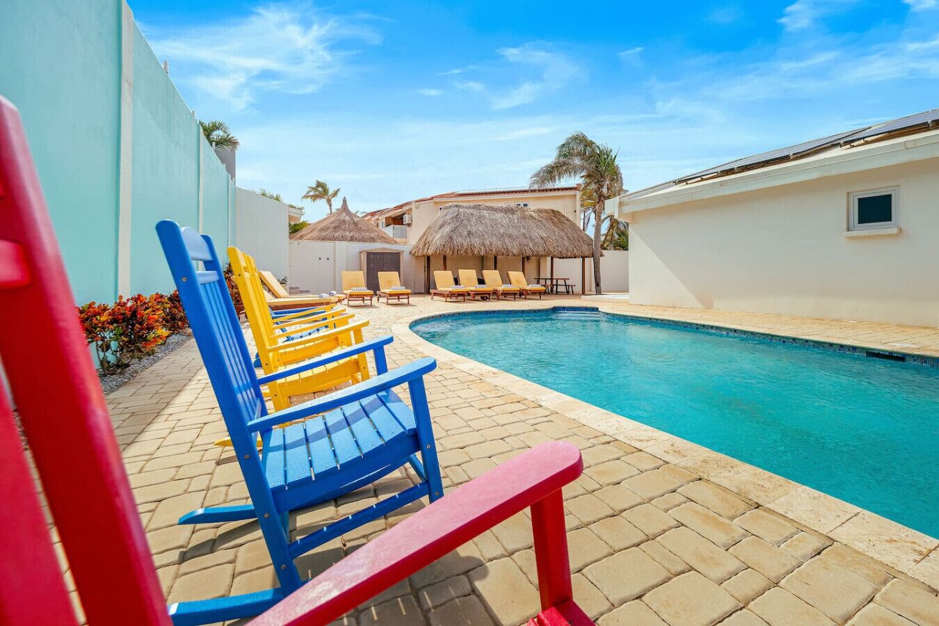 Ein Poolbereich mit blauen und roten Liegestühlen, einer Struktur mit Reetdach und einem klaren, blauen Pool.