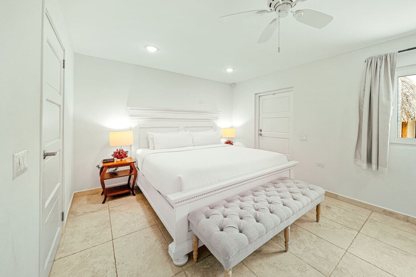 Un dormitorio blanco con cama, mesitas de noche, un banco, un ventilador de techo y una puerta.