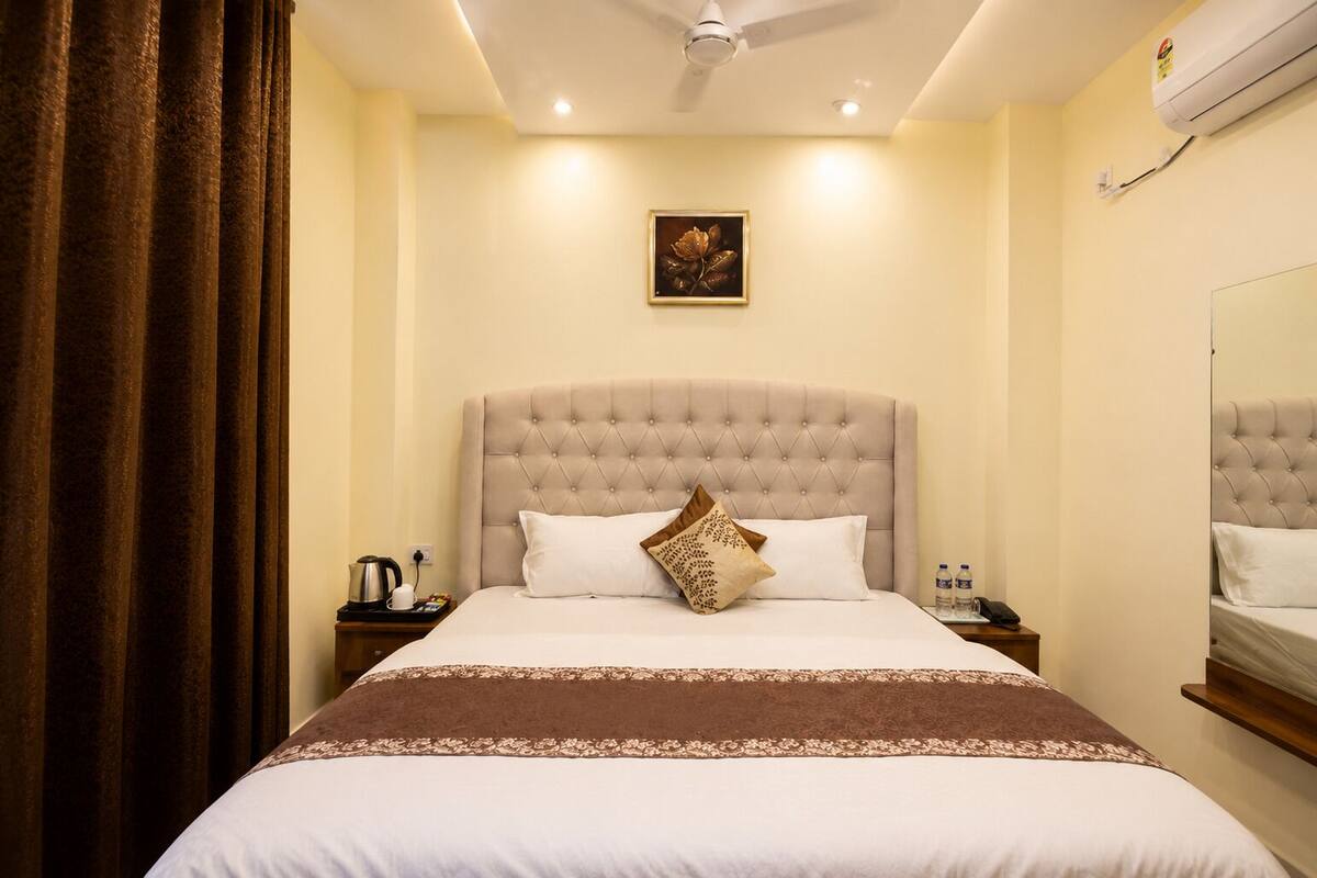 Quarto de hotel com cabeceira estofada, cama com roupa de cama branca e castanha, mesa de cabeceira com chaleira e chávena, e ar condicionado fixado na parede.