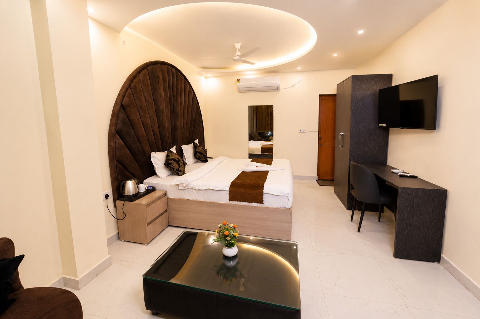 A modern hotel room with a bed, bedside table, TV, desk, and a glass coffee table.