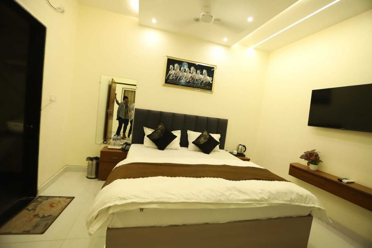 A modern hotel bedroom with a large bed, a flat-screen TV, a framed picture on the wall, and a small plant on a nightstand.