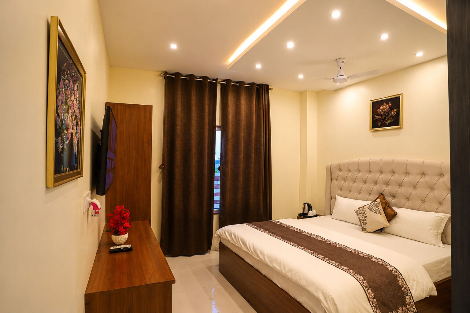 A hotel room with a bed, a wooden nightstand, a TV, a ceiling fan, and a window with curtains.