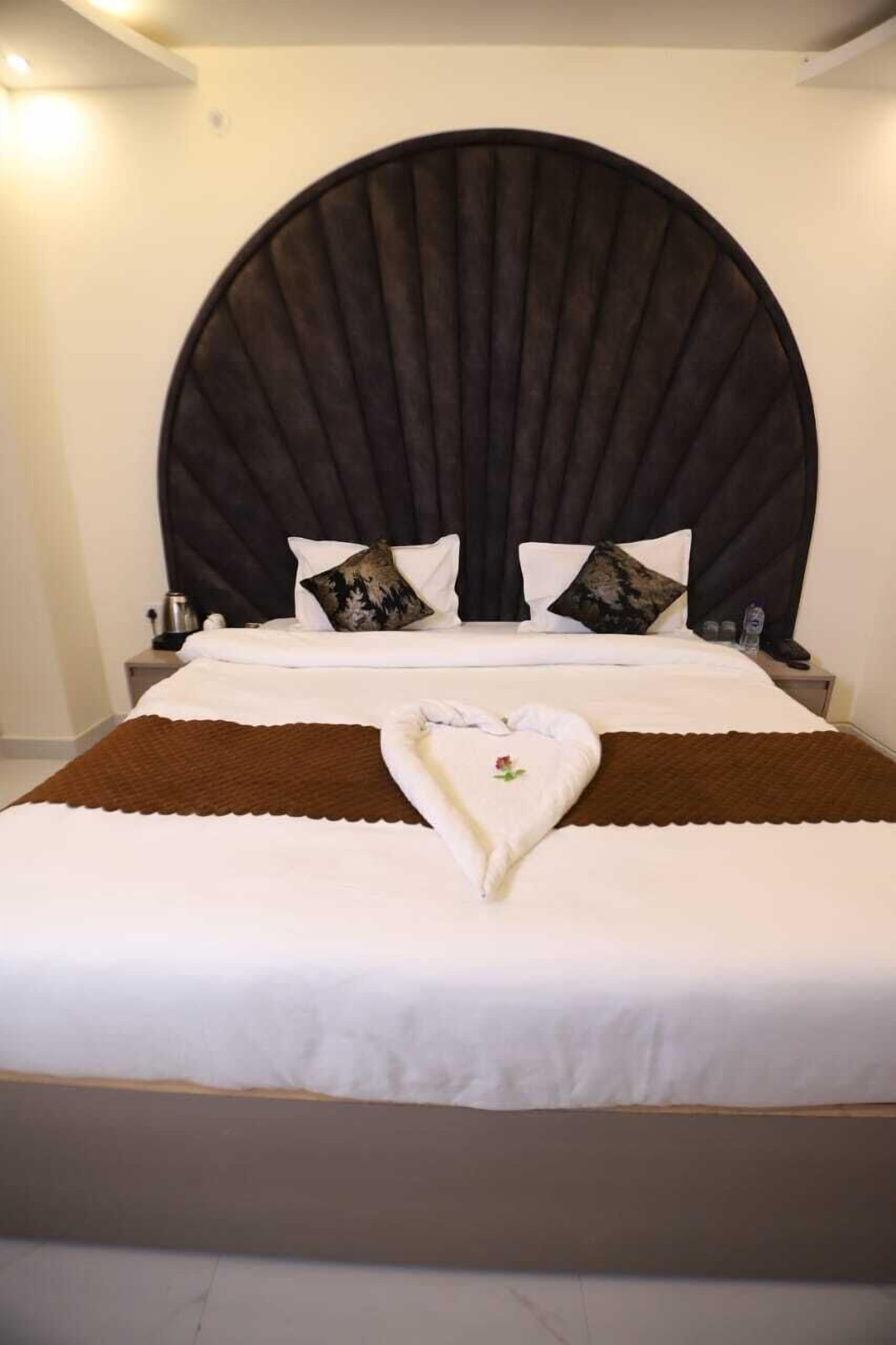 Un lit bien fait, agrémenté d’une serviette en forme de cœur, d’une grande tête de lit sombre et de deux tables de chevet avec des lampes.