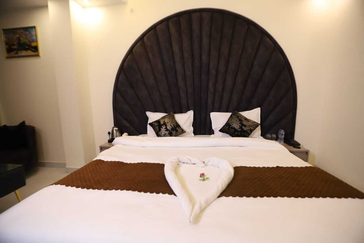 Habitación de hotel con una cama grande, cabecera oscura, ropa de cama blanca y una toalla con forma de corazón sobre la cama.