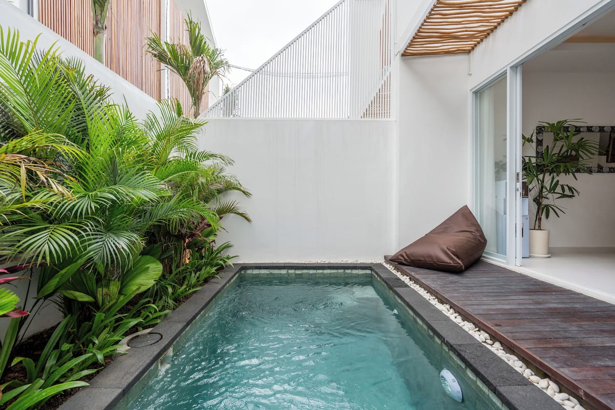 A small, rectangular swimming pool with a wooden deck and white pebbles around the edges, surrounded by lush green tropical plants.