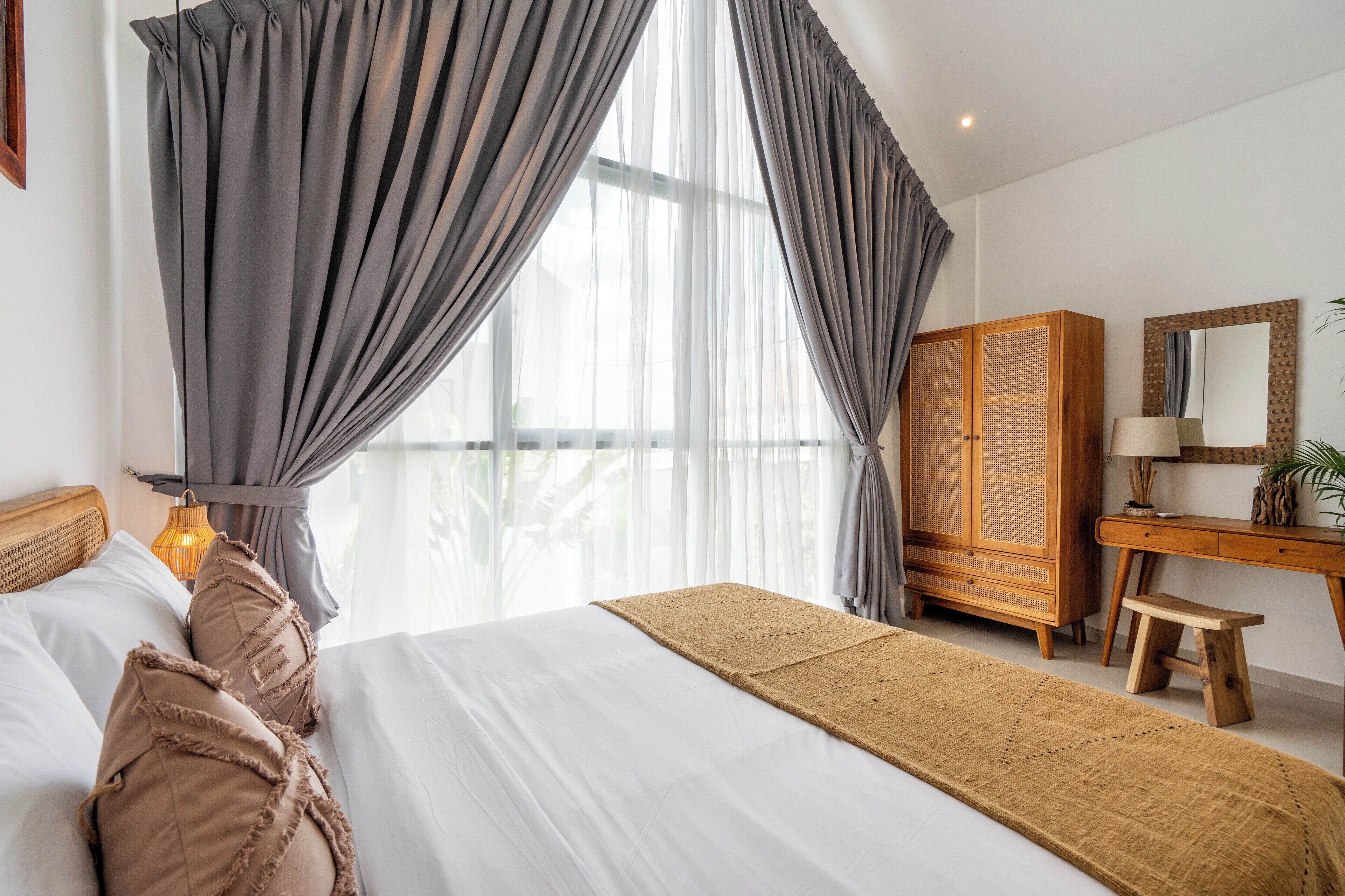A bedroom with a bed, grey curtains, a wooden wardrobe, a desk with a lamp, and a view of the outdoors.