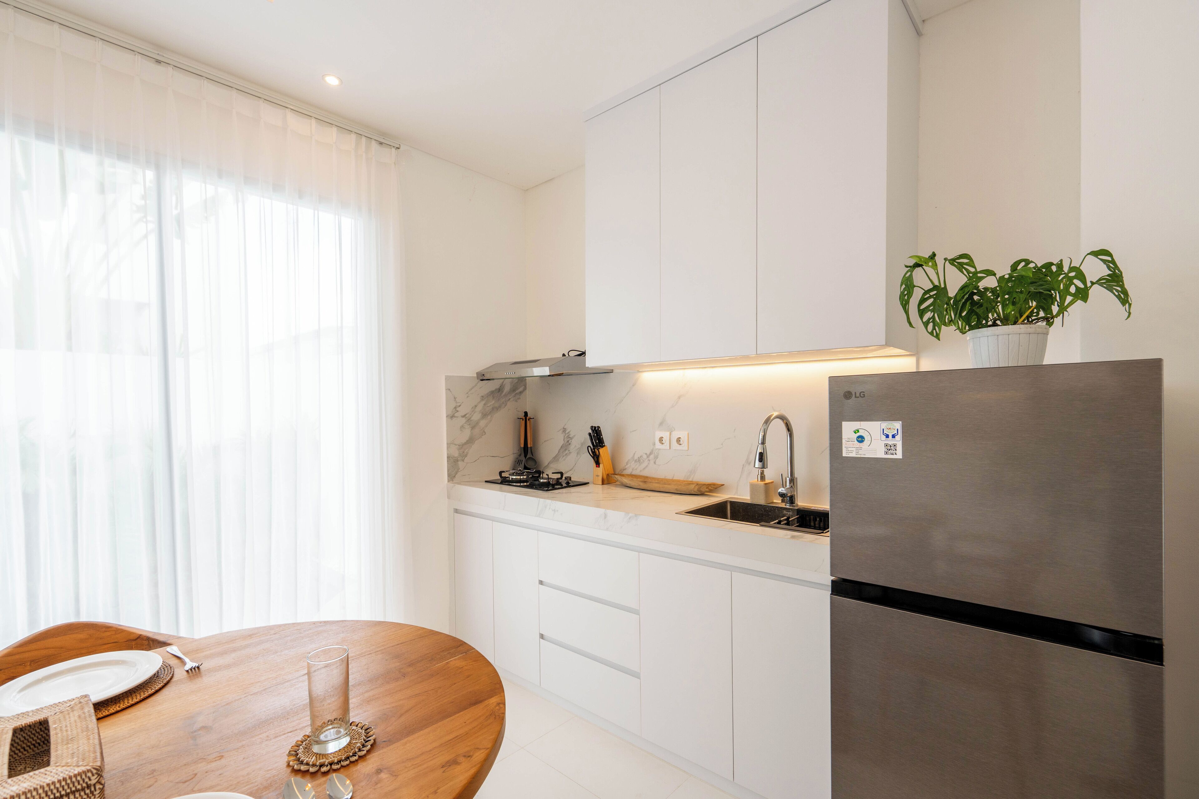 A modern kitchen with white cabinetry, a stainless steel refrigerator, a wooden dining table, and a potted plant.