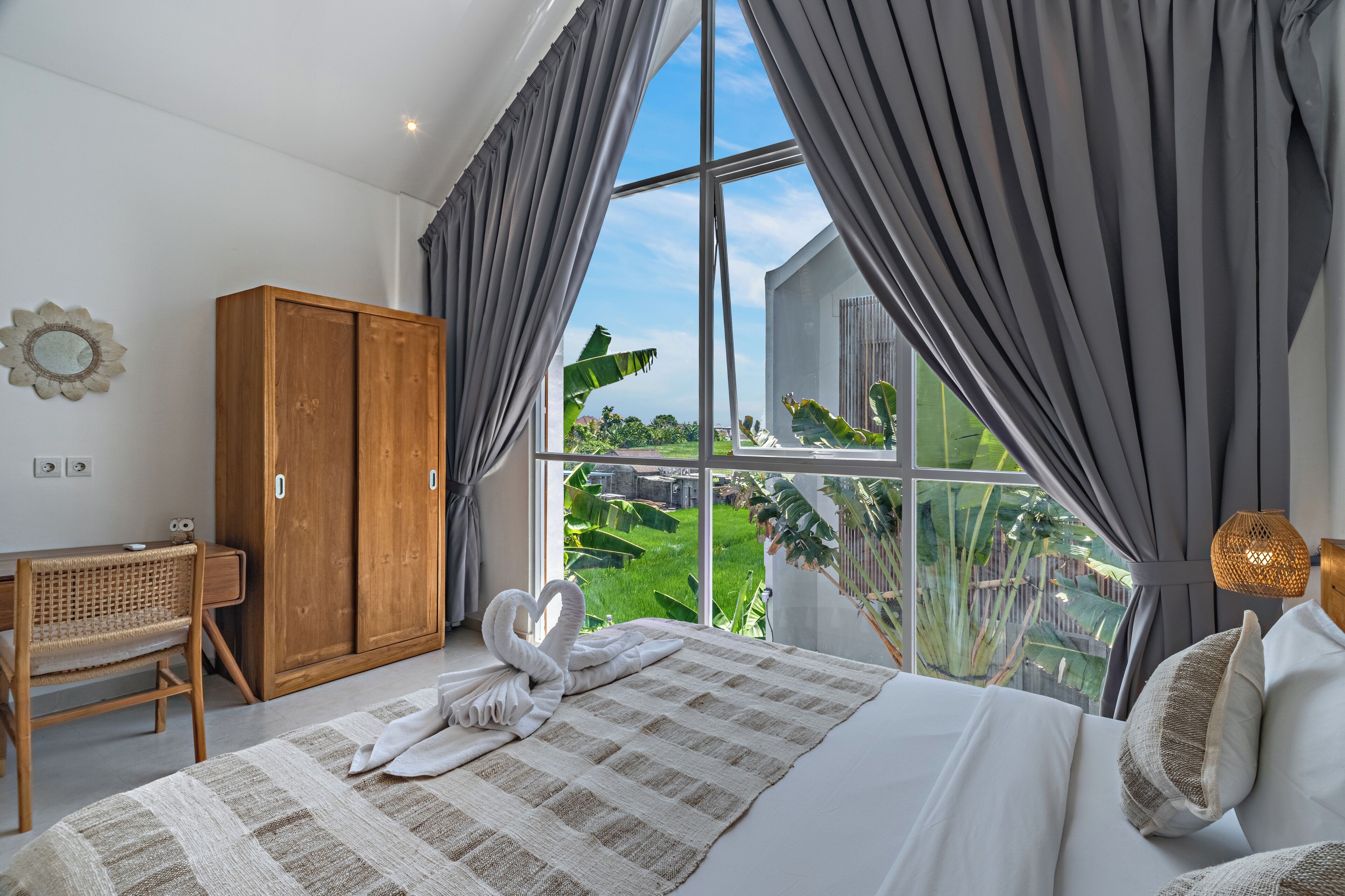 Ein Schlafzimmer mit großem Fenster, einem Holzschrank, einem Bett mit handtuchförmigem Schwan und einem Schreibtisch mit Korbstuhl.