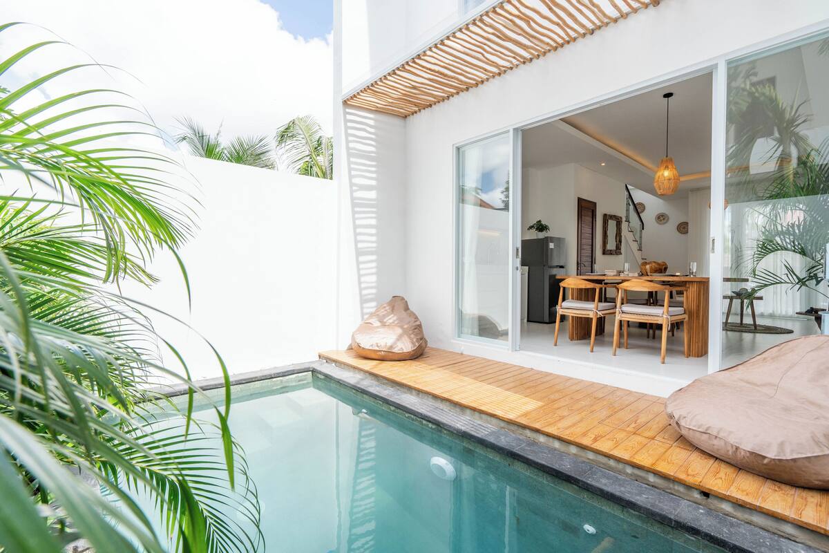 A modern poolside area with a glass door leading to a dining area, surrounded by lush greenery.