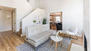 A spacious living room with a grey sofa, a small white table, and a blue patterned rug. A staircase leads to an upper level, and a kitchen is visible in the background.