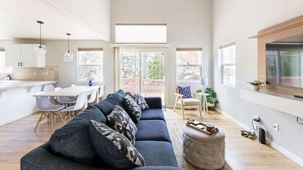 A modern living room with a dark blue sofa, a dining area with a white table and chairs, and a kitchen with white cabinets.