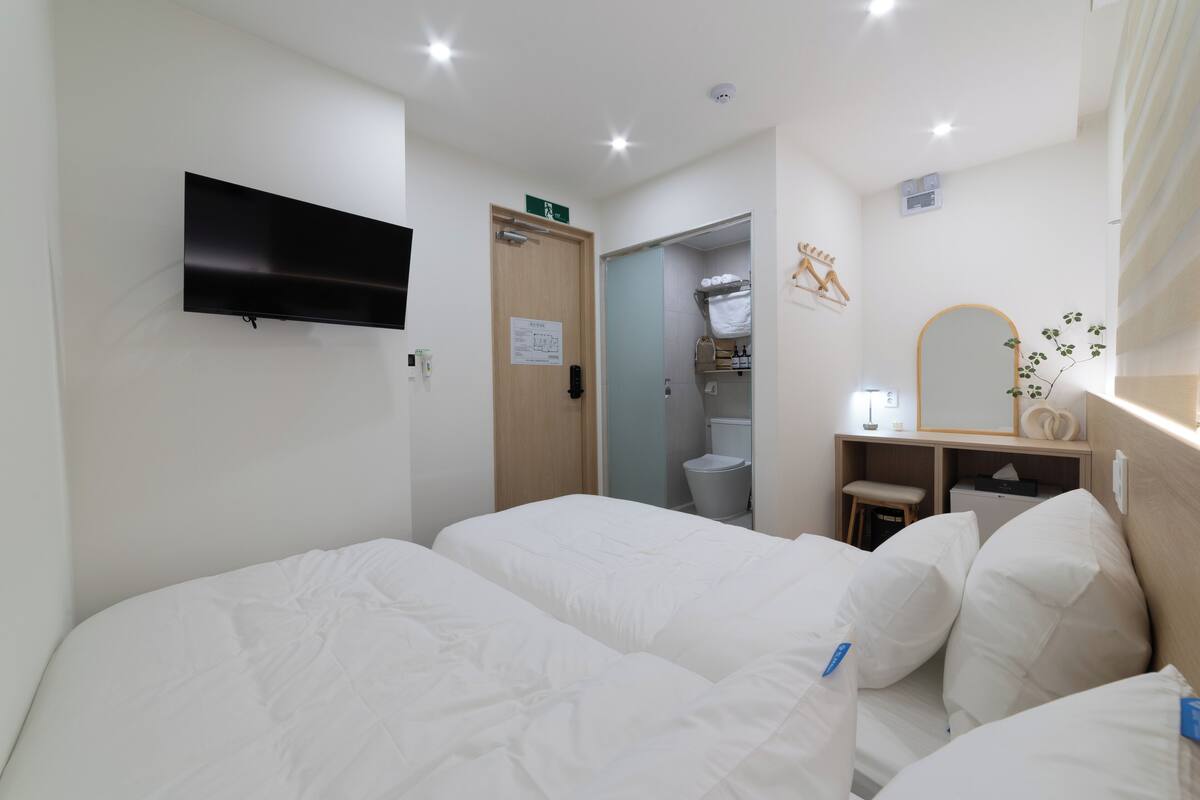 Habitación de hotel con dos camas, televisor, espejo, escritorio y baño visible a través de una puerta abierta.
