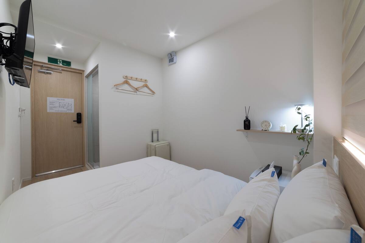 Habitación de hotel con una cama, una mesita de noche, un espejo, un perchero y un pequeño estante con objetos decorativos.