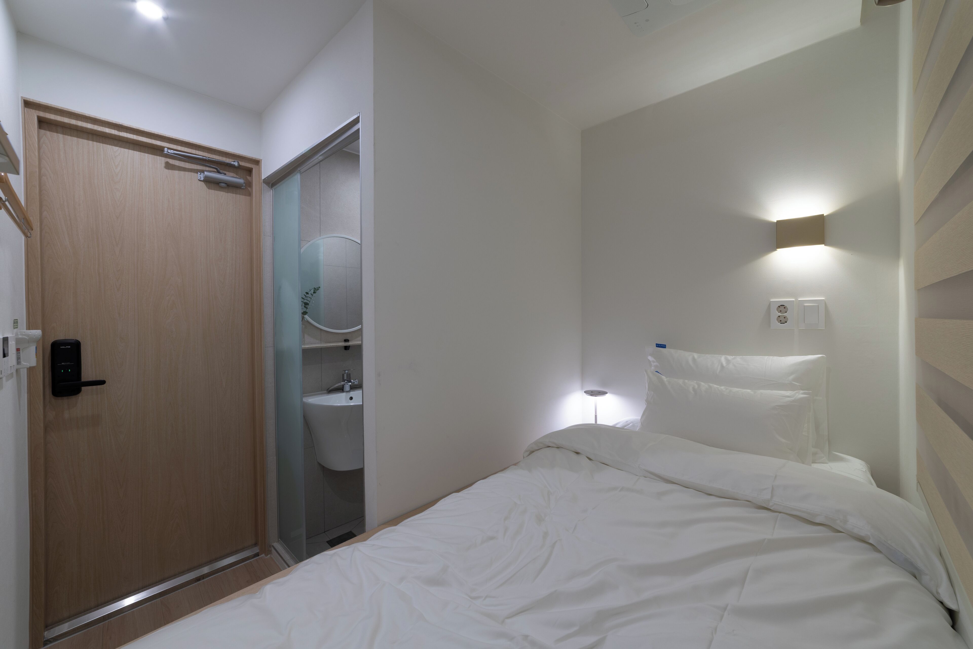 Un dormitorio con puerta de madera, un baño con inodoro y lavamanos, y una lámpara de noche.