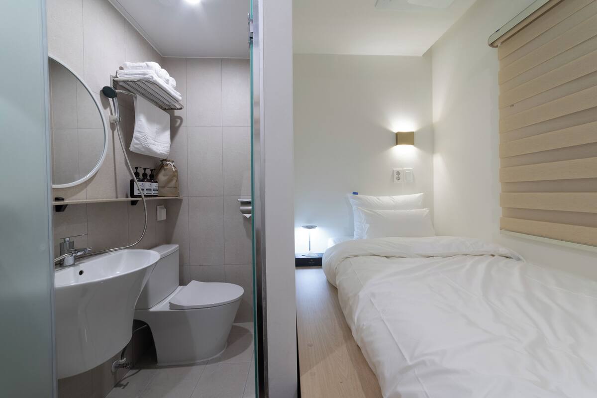 Un baño moderno con lavamanos, inodoro y ducha. Hay una cama con ropa de cama blanca y una mesita de noche.