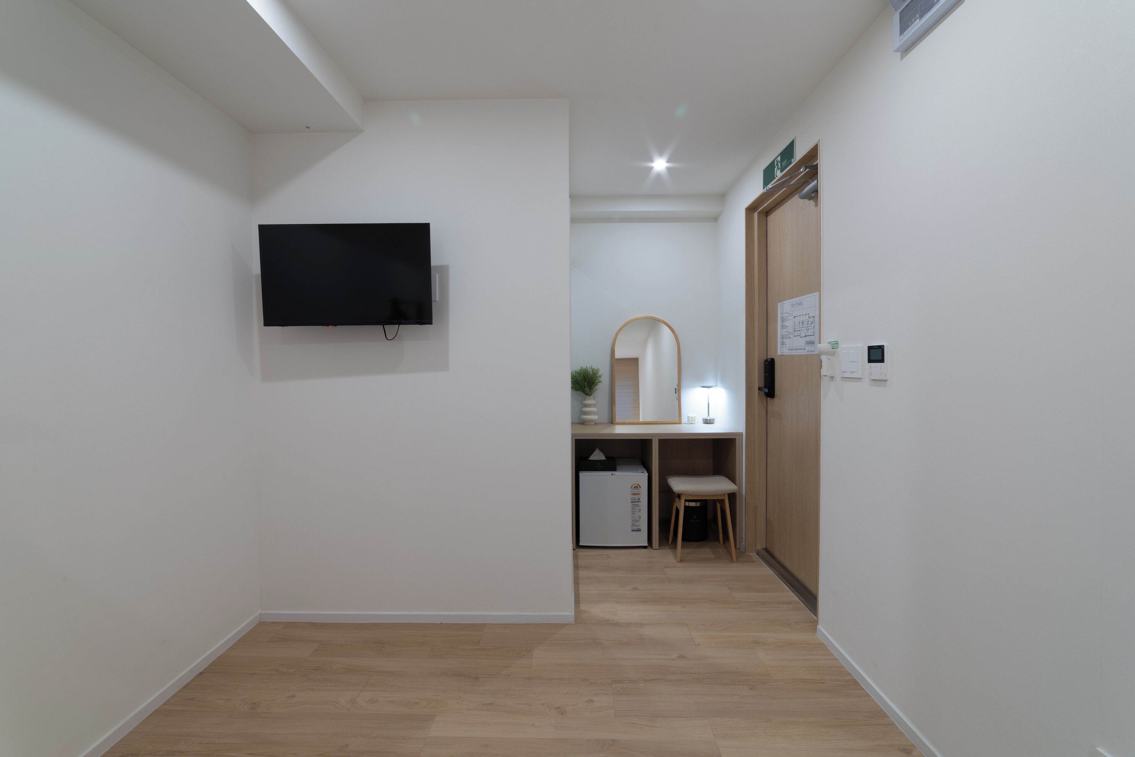 A hallway with a wooden floor, a flat-screen TV mounted on the wall, a desk with a chair, a mirror, and a small refrigerator.