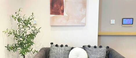 Un sofá gris con dos cojines estampados y un cojín blanco redondo, un cuadro abstracto grande, una planta pequeña y una pantalla digital en la pared.