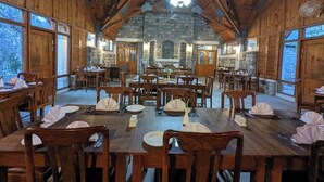 A spacious dining area with wooden walls and a stone fireplace. Tables are set with white tablecloths and silverware. - Destination Hotel Khaplu (Khaplu)