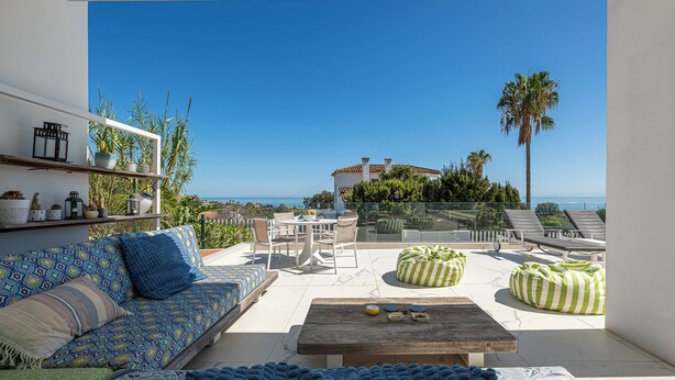 A spacious balcony with a sofa, chairs, and a coffee table. There are two bean bags and a glass railing. The view includes a palm tree and a building across the water. - Heated Pool Villa / Sea & Gibraltar View (Torreguadiaro)