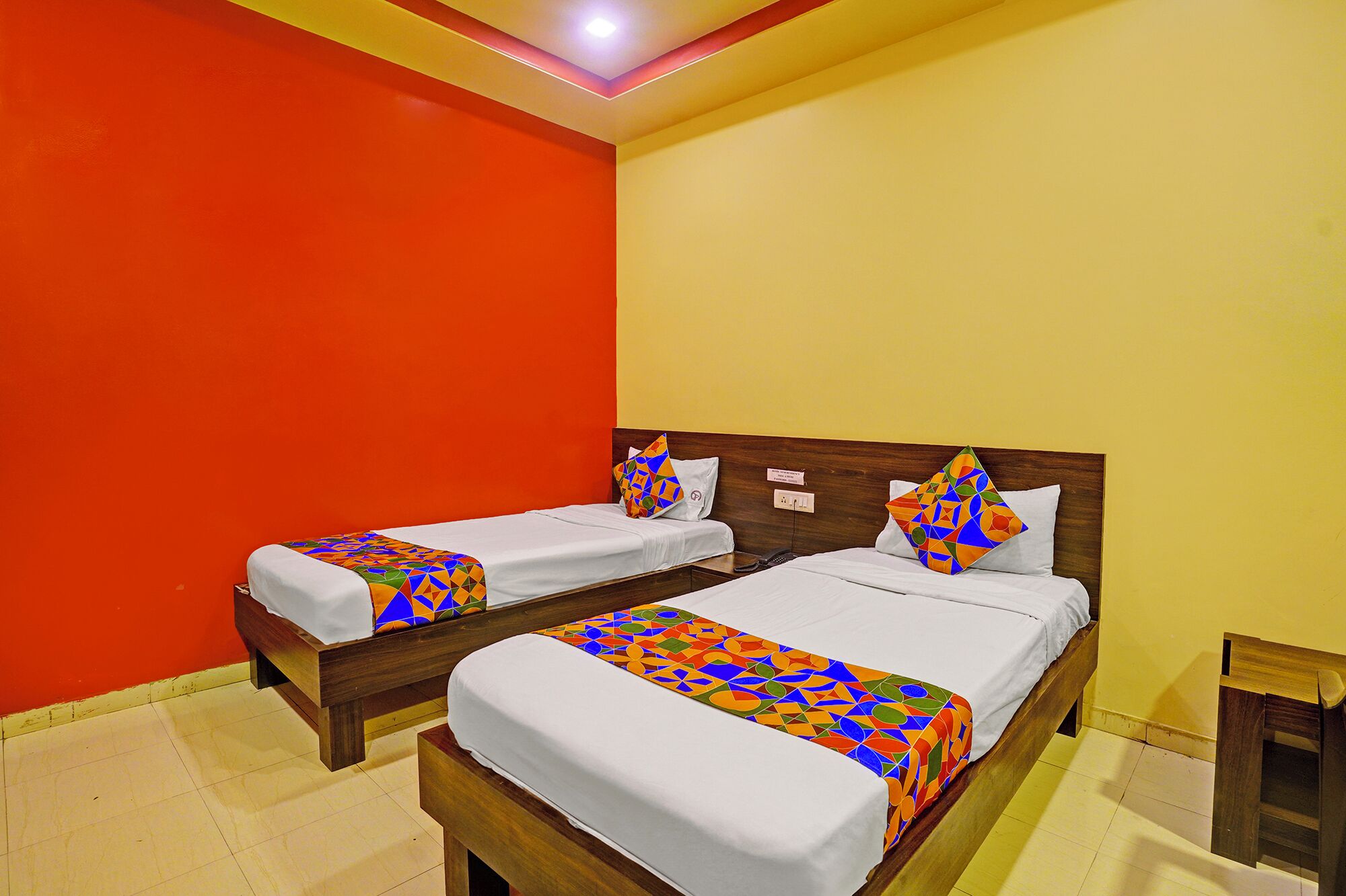 A hotel room with two beds, each with a colorful geometric-patterned bedspread and matching pillows. The room has a wooden headboard, a small bedside table, and a nightstand.
