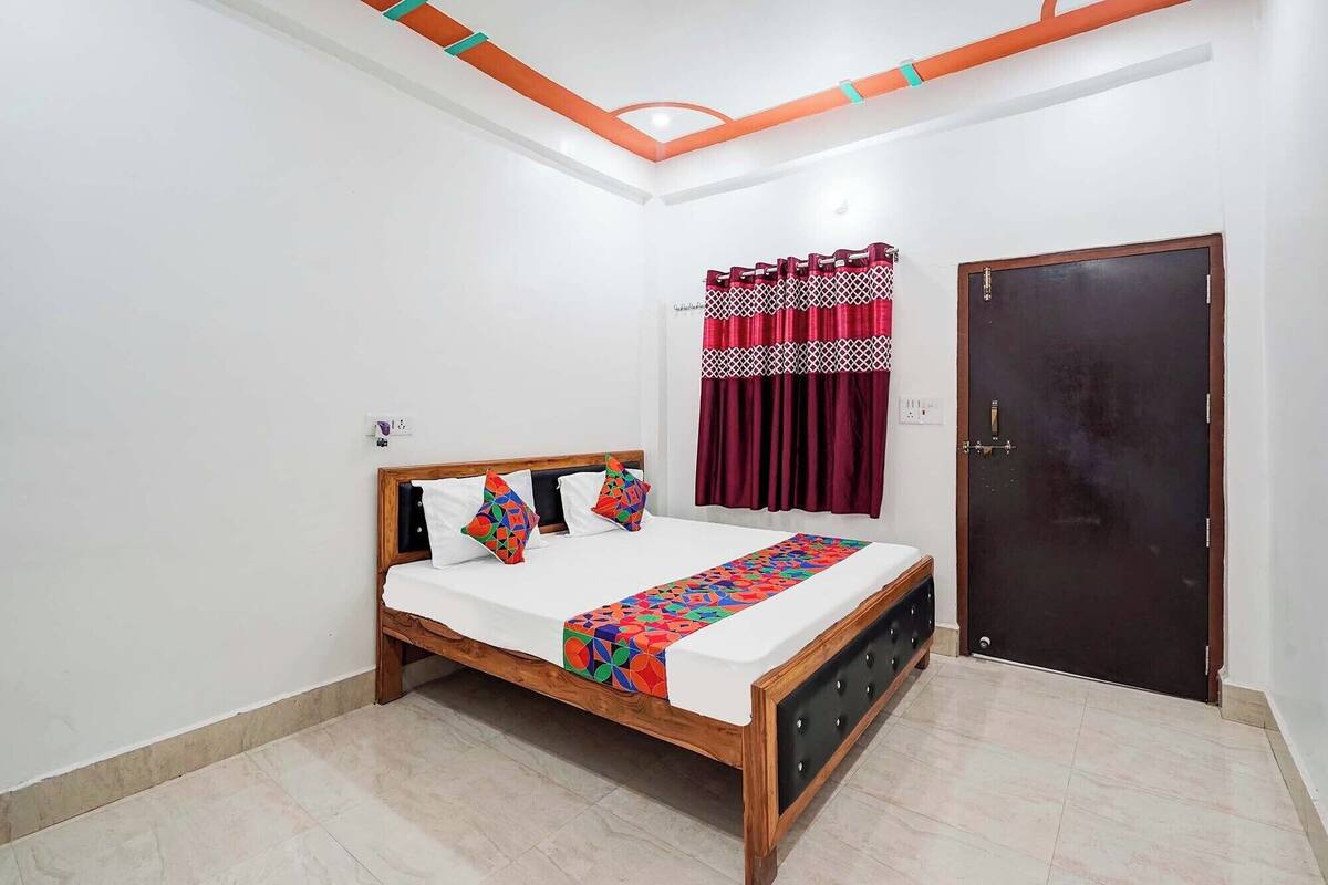 Un dormitorio con cama de madera, ropa de cama blanca, colcha colorida, puerta oscura y techo blanco con detalles anaranjados.
