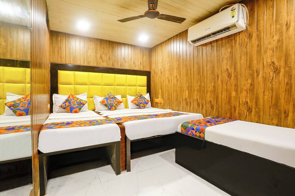 A hotel room with a wooden paneled wall, a ceiling fan, an air conditioning unit, a bed with a yellow headboard, and a bedside table.