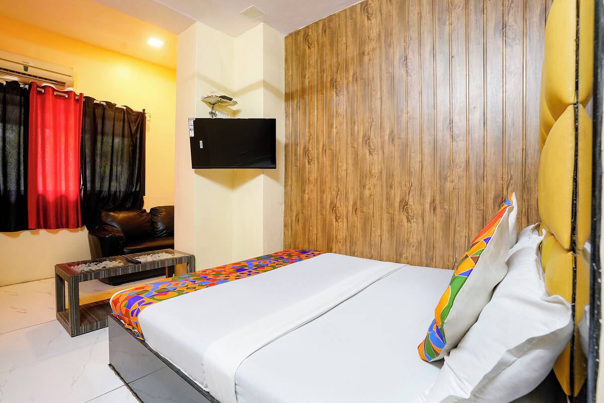 A hotel room with a bed, a wooden headboard, a TV mounted on the wall, a window with curtains, and a small table with a tray of snacks.