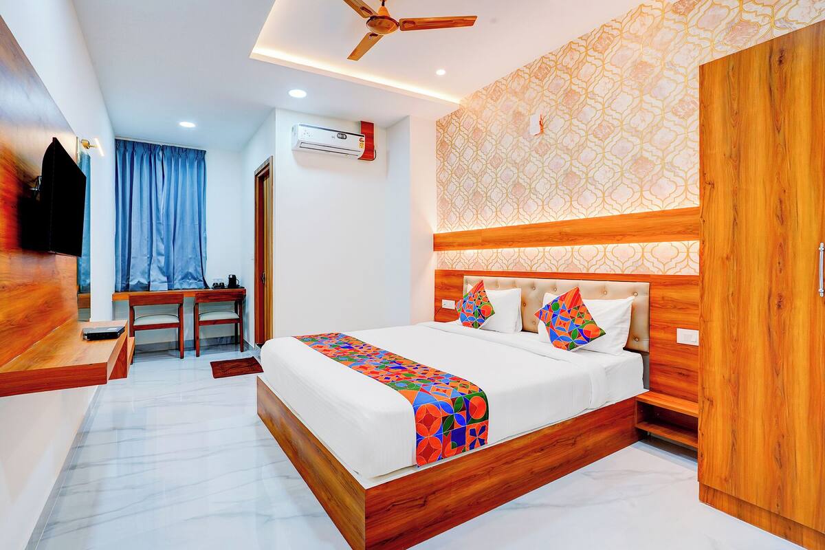 A hotel room with a bed, wooden headboard, a desk with a chair, a TV, and an air conditioning unit.