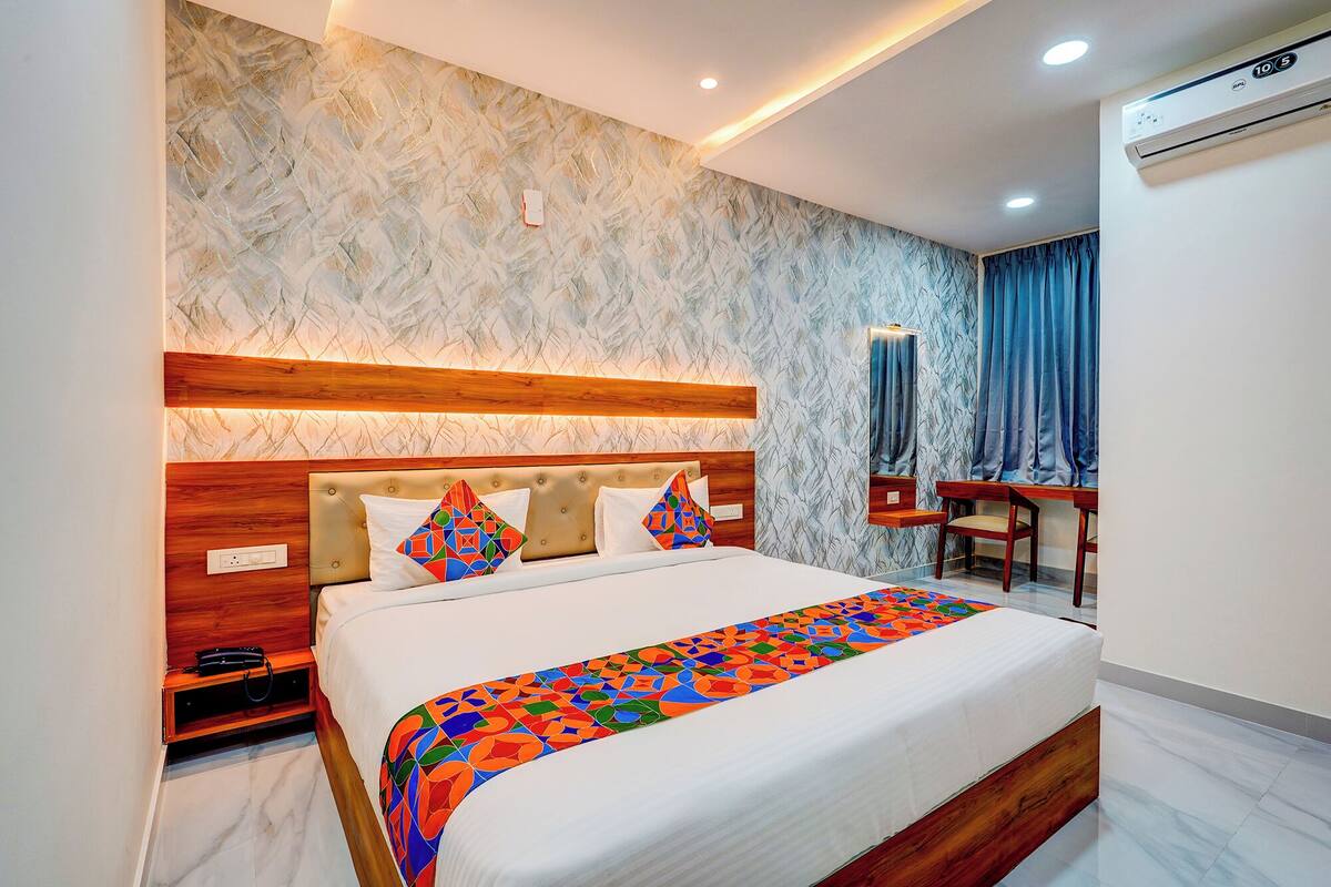 A modern hotel room with a large bed, a wooden headboard, a desk, and an air conditioning unit.
