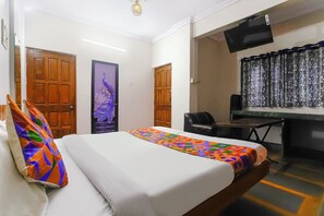 A hotel room with a bed, a desk, and a chair. There are two doors, one with a peacock design and another with a wooden panel. A TV is mounted on the wall above the desk.
