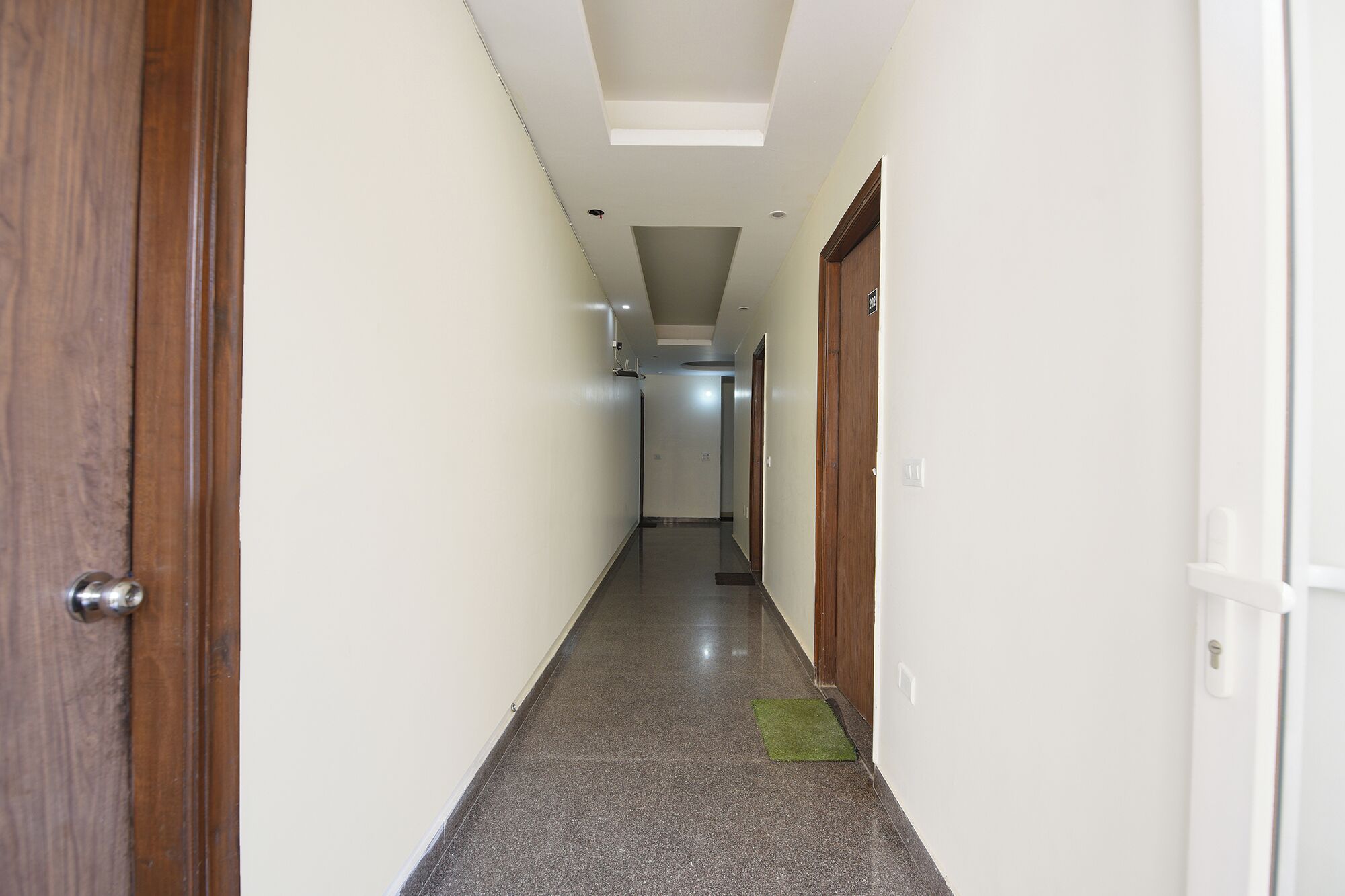 A narrow hallway with a wooden door on the left, a green mat at the end, and a white door on the right.