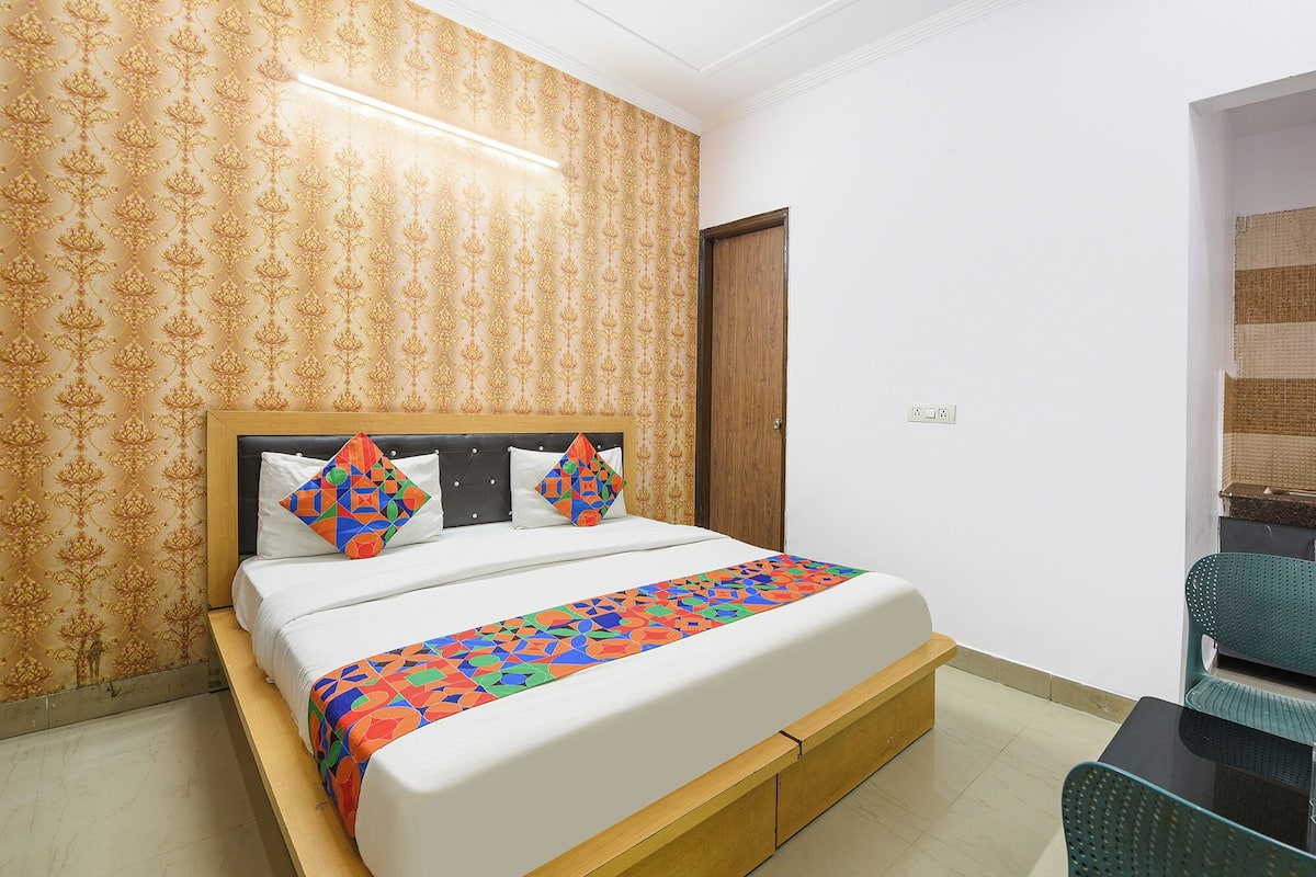 A neatly made bed with a colorful geometric-patterned bedspread, a wooden headboard, and matching pillows. The room has patterned wallpaper and a wooden door.