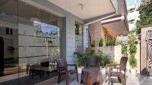 An outdoor seating area with wicker furniture, potted plants, and a glass wall reflecting the surroundings. - Fabhotel The Flora (Gurugram)
