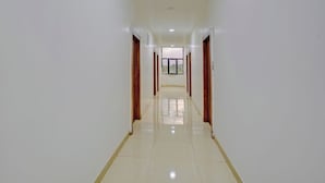 A long, narrow hallway with white walls and wooden doors, leading to a room with a large window. - Fabhotel Ashiyana (Pune)