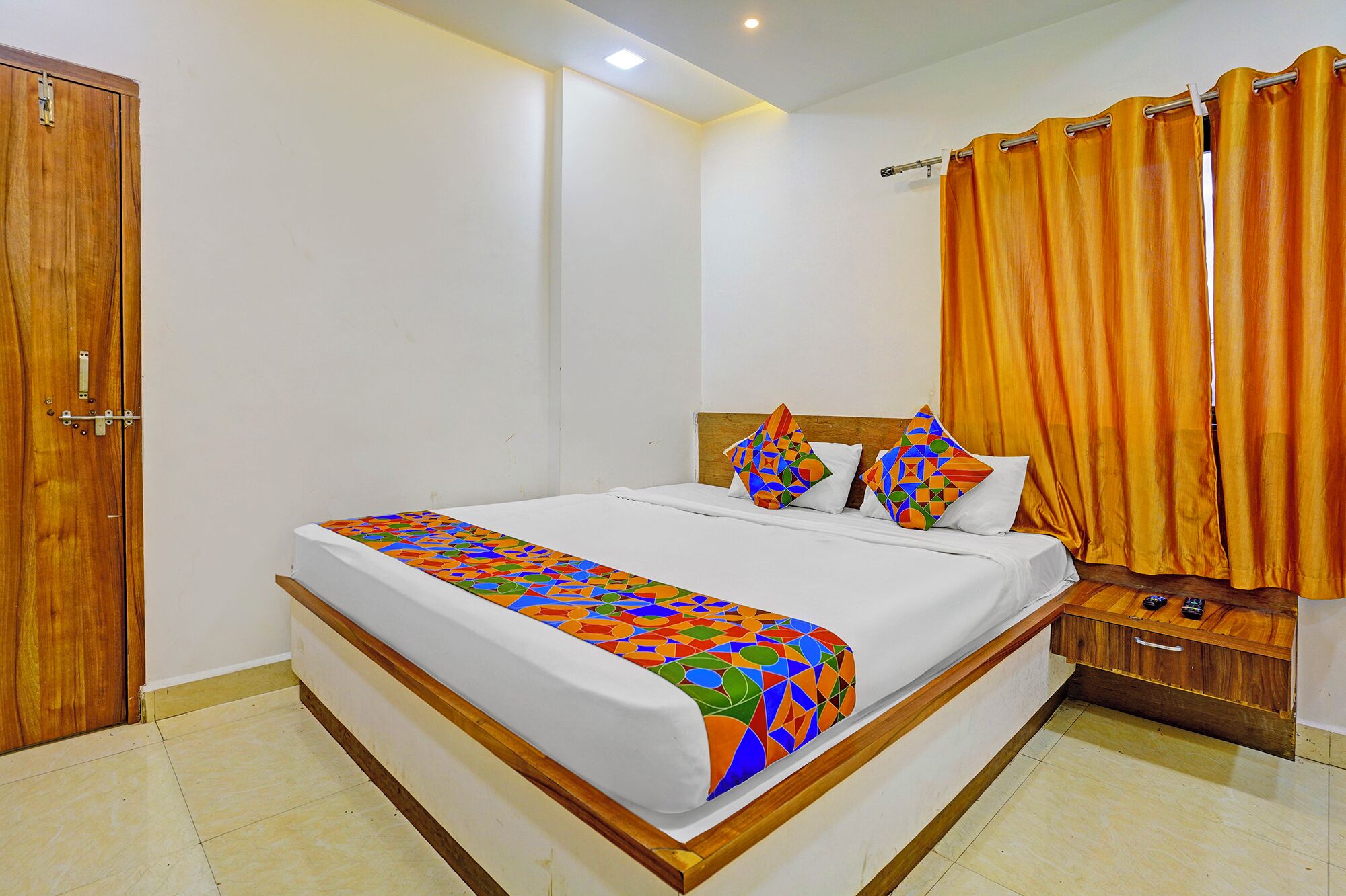 A bedroom with a wooden bed frame, white bedding, colorful geometric-patterned throw, and a wooden nightstand.