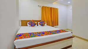 A neatly made bed with a colorful geometric-patterned bedspread, a wooden headboard, and a window with orange curtains. - Fabhotel Ashiyana (Pune)
