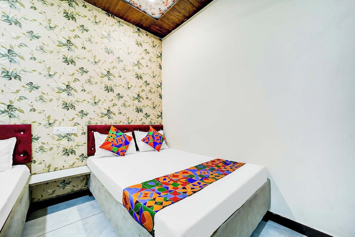 A small, neatly arranged bedroom with a floral wallpaper, a wooden ceiling, and a bed with colorful geometric pillows and a matching bedspread.