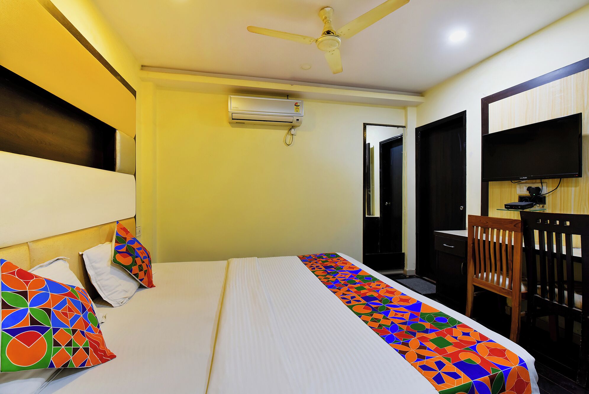 A hotel room with a bed, a desk, a chair, an air conditioning unit, and a television.