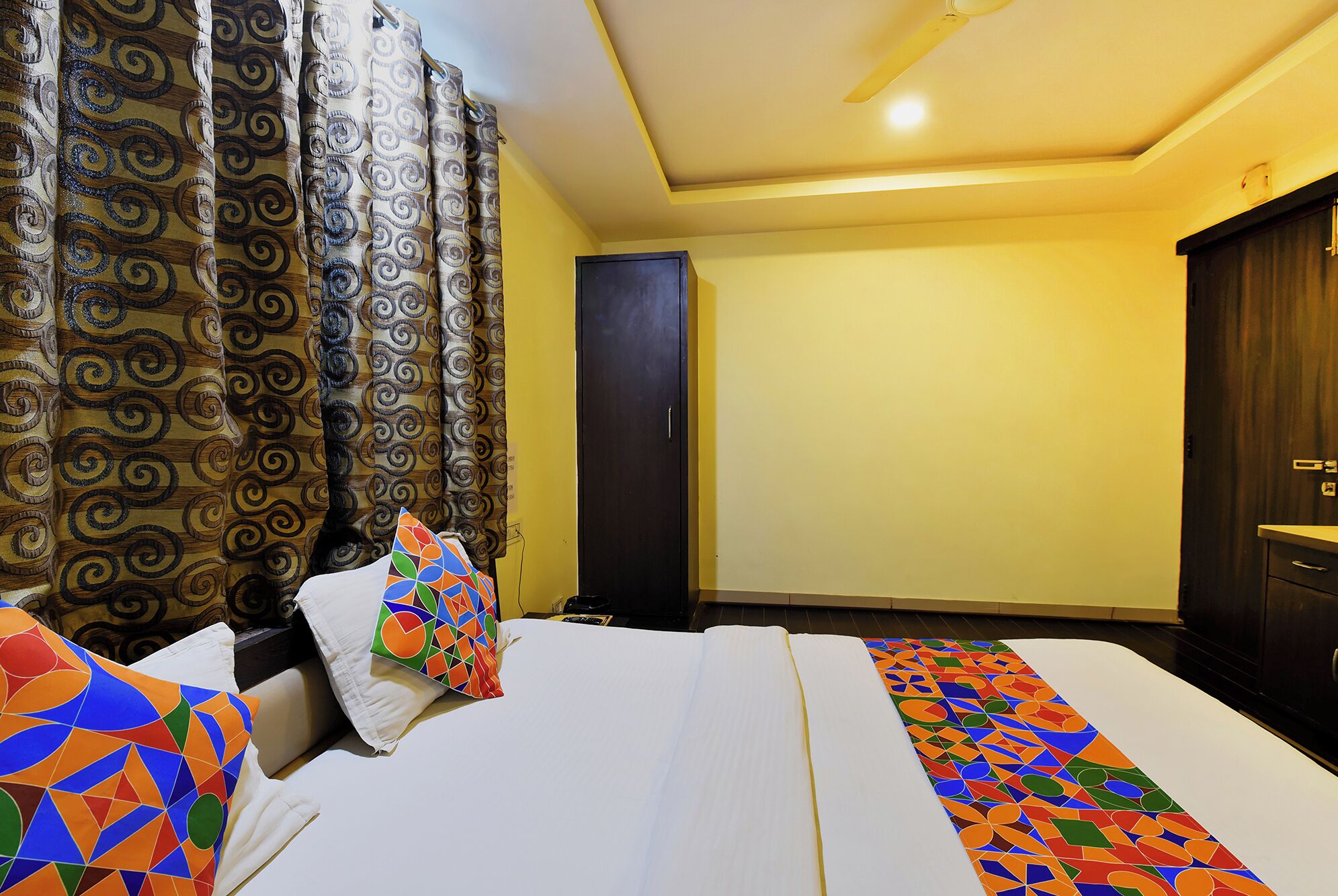 A hotel room with a bed, curtains, a wardrobe, and a pillow with a colorful geometric pattern.