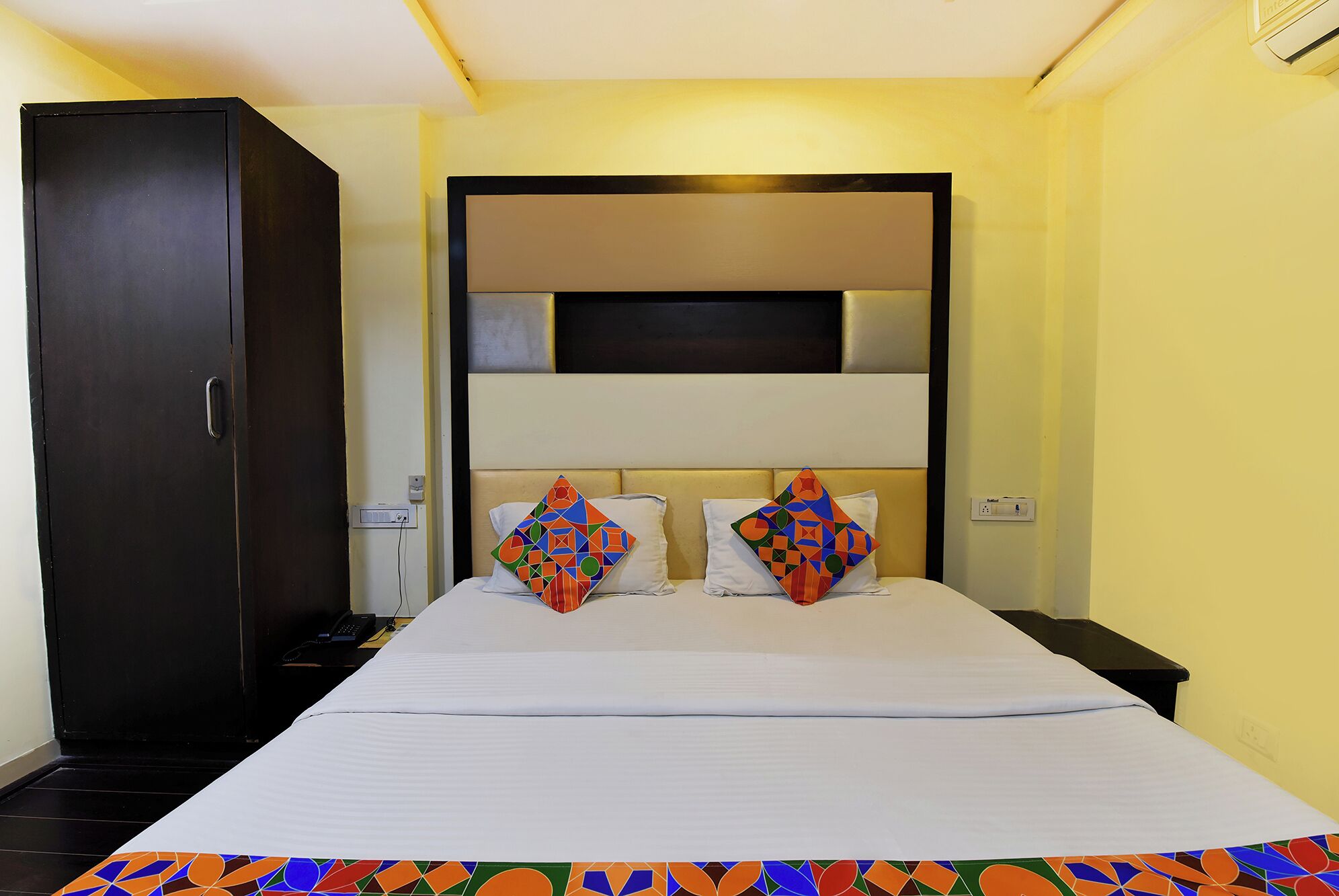 A hotel room with a large bed, two pillows with geometric patterns, a wardrobe, and a bedside table.