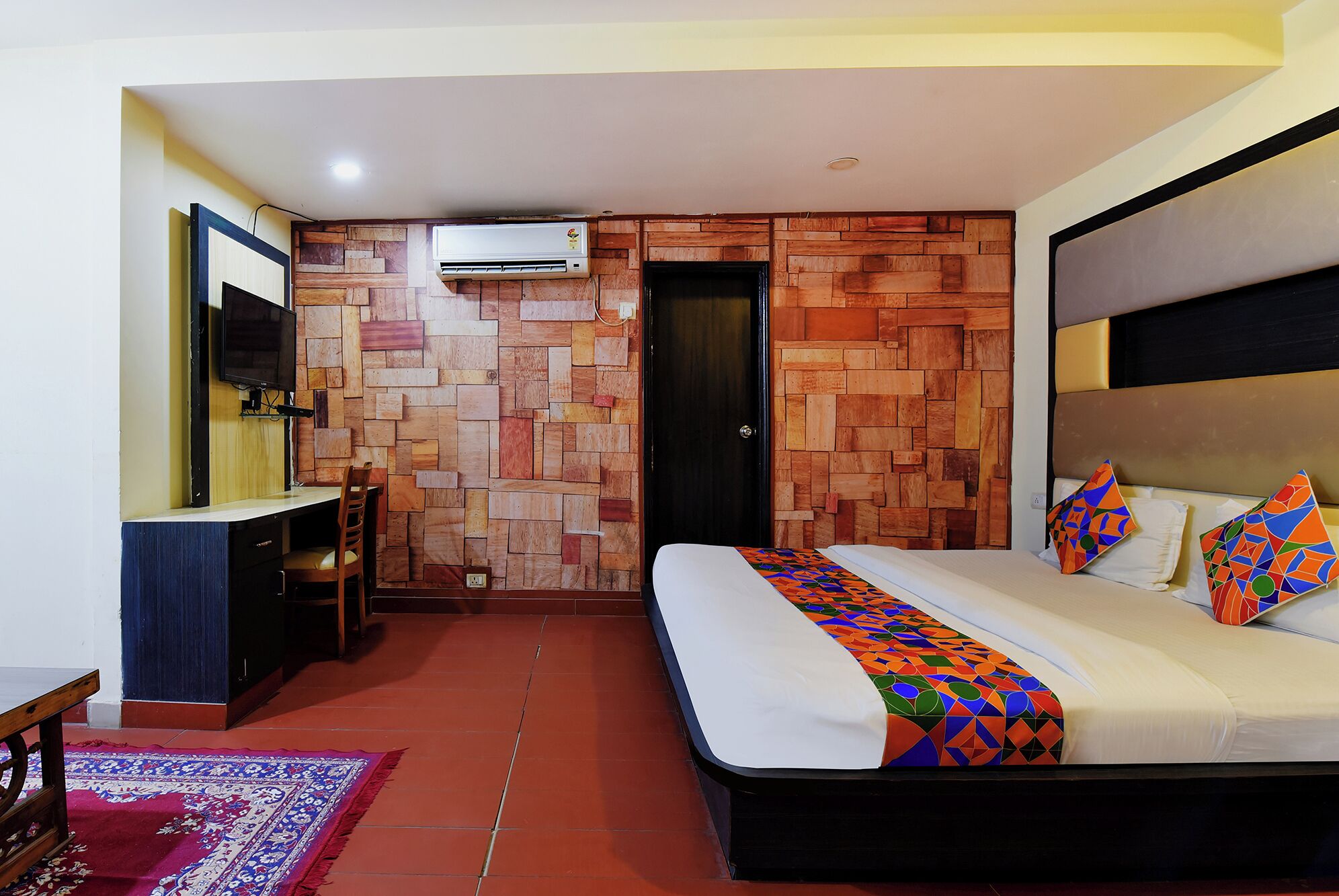 A hotel room with a wooden wall design, a bed with colorful pillows, a TV mounted on the wall, and a desk with a chair.
