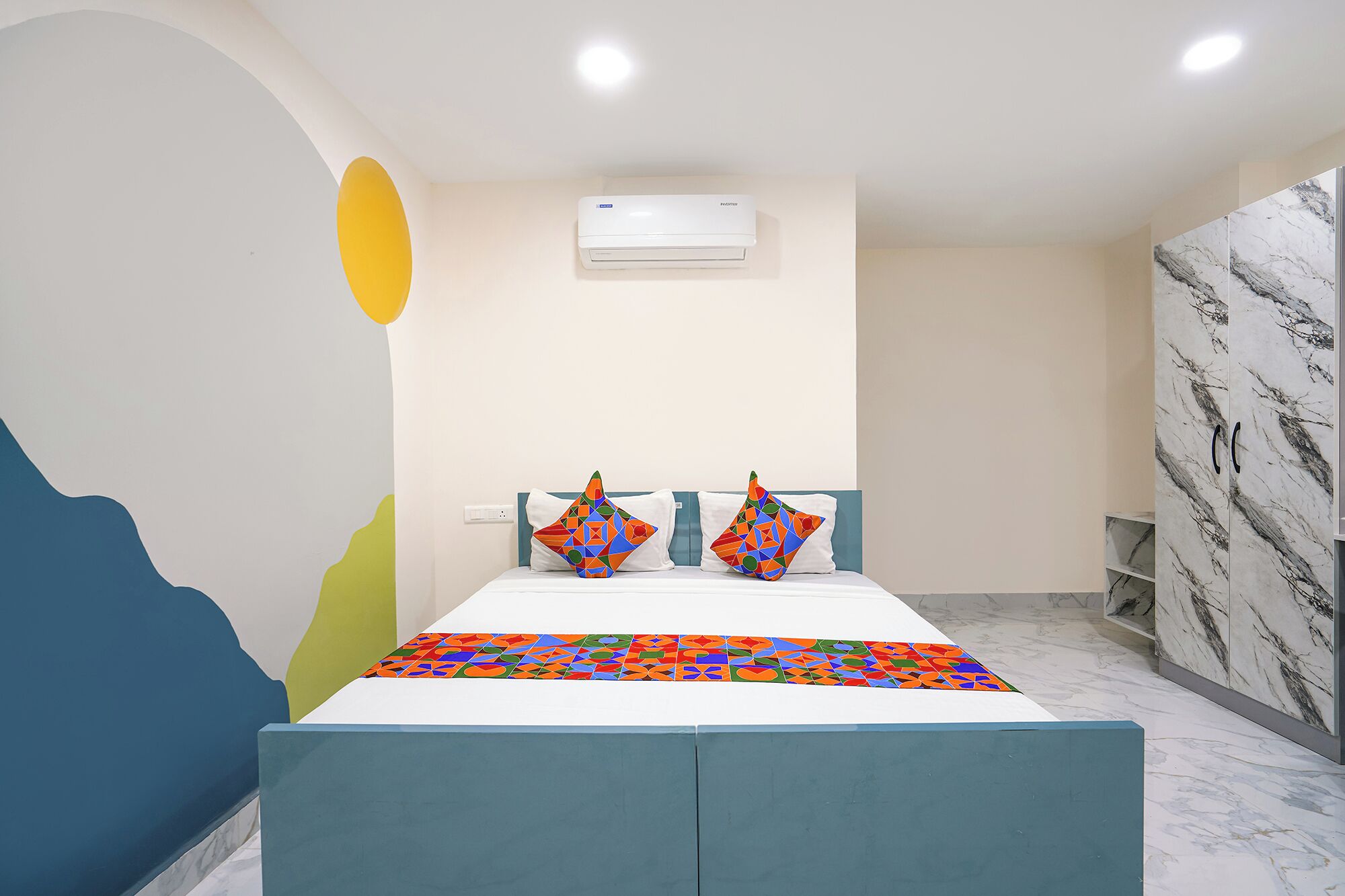 Hotelkamer met een bed, kleurrijke kussens, een wandairconditioner en een wand met een marmerlook.