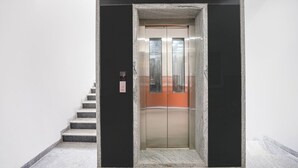 A modern elevator with a metallic interior and a glass panel, flanked by a staircase with stone steps and a black frame.
