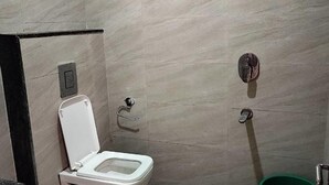 Une petite salle de bain carrelée, équipée de toilettes, d’un sèche-mains et d’une poubelle verte.
